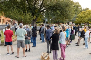 People are standing around on gravel under tall trees looking in the direction of a speaker.