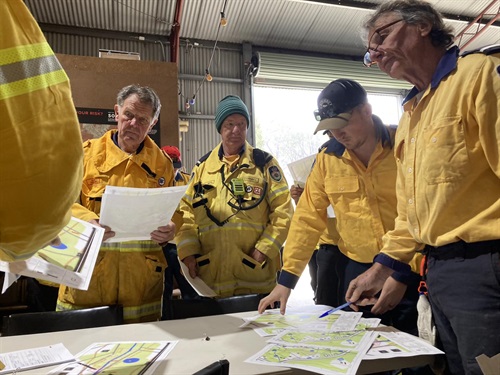 Five men in yellow NSW RFS uniforms standing in a fire shed around a table looking at fire maps