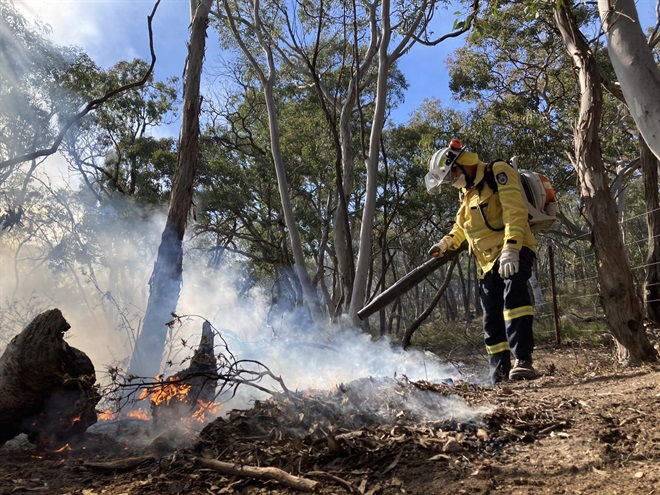 NSW RFS volunteer in yellow clothing using a blower to increase flames in leaf litter that is making smoke