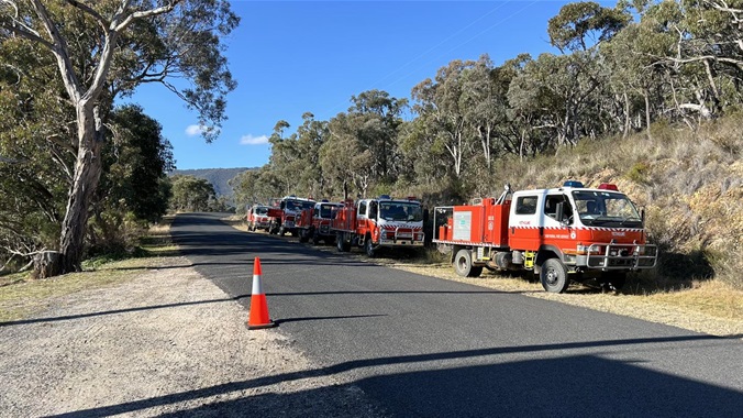 Five NSW RFS red trucks parked alongside a dark road with tall eucalyptus trees bound either side of the road